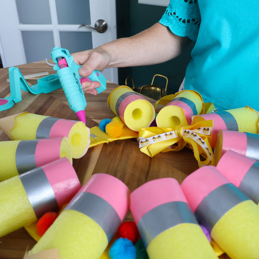 Colorful pool noodle crafts on a table with a person using a Lynn Lilly glue gun.